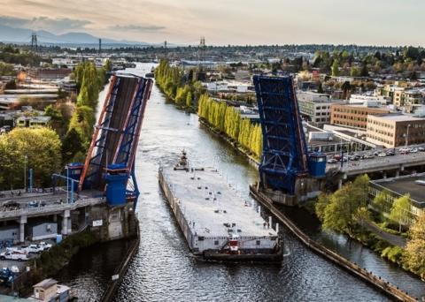 A tugboat guide a large concrete pontoon on the water underneath the raised roadways of the Freemont Bridge.