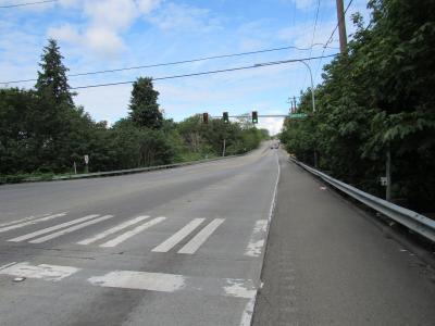 State route 900 stretches from the foreground straight ahead showing the westbound shoulder where a new sidewalk will be built at 68th Avenue South. The sky is blue with white puffy clouds and greenery borders both sides of the roadway. The are no vehicles or people visible in the image.