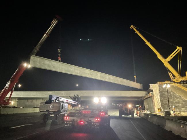 Two cranes lifting a long piece of concrete that is part of a new bridge. Bright lights illuminate the construction site, creating a stark contrast to the dark night sky. Large construction vehicles sit on the road, slightly underneath the concrete slab.