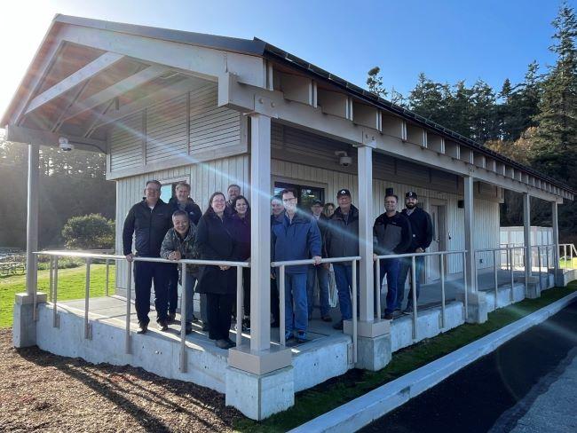 The image shows a group of people standing on the porch of a long, narrow building with a sloped roof.