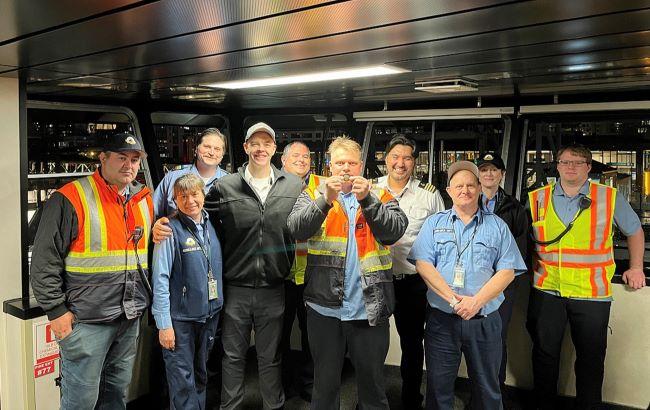 A group of ten people standing together inside a well-lit interior setting. Several individuals wear orange reflective safety vests over casual clothing, while others wear light blue shirts. The person in the middle is in a dark jacket and cap, smiling with an arm around others.