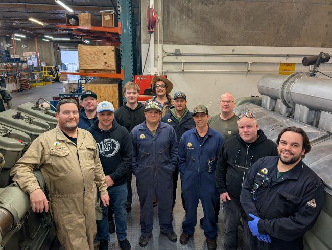 11 people in an industrial workshop, standing in front of engine equipment