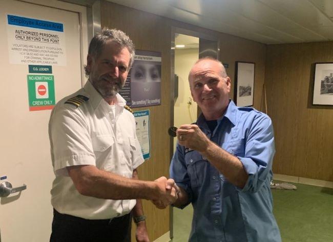 Two people shaking hands in a ship's corridor with one holding up a coin