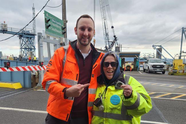 Two people in high-visibility jackets at a ferry terminal with cranes and vehicles in the background and one holding up a coin