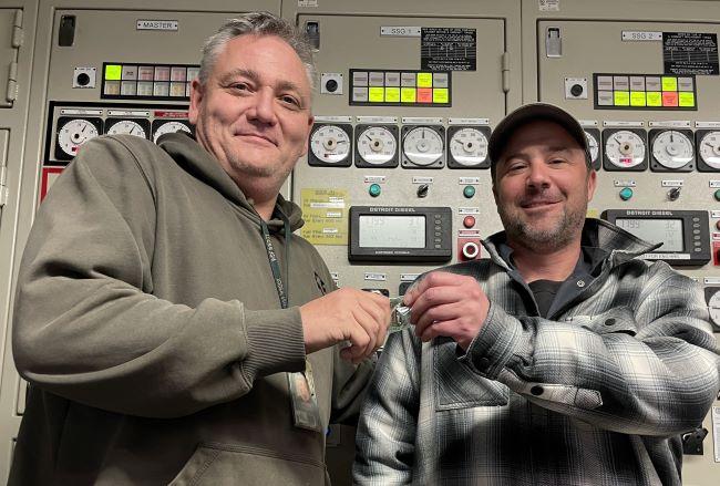 Two people smiling in front of an engine room control panel with both holding up a single coin