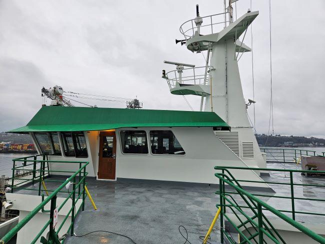 Top deck of a ferry with green-roofed bridge, large windows and green railings against a cloudy sky