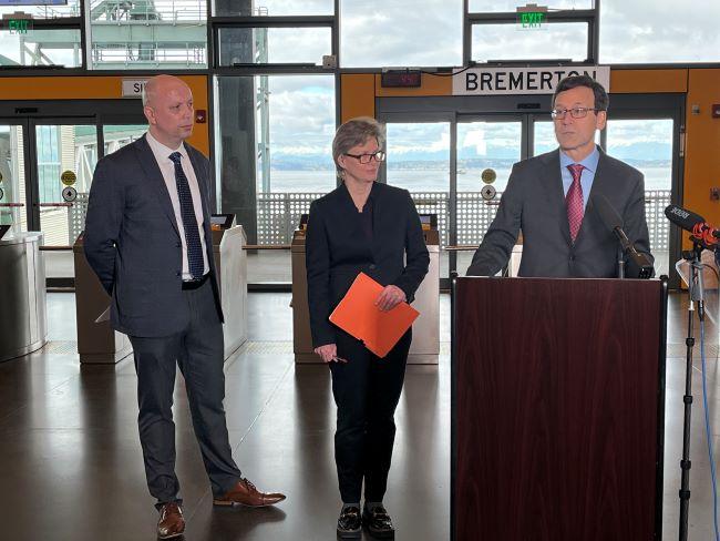 Three people standing inside Colman Dock terminal building with one speaking at a podium